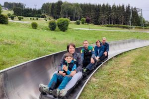Familie in einem Sommerrodel umgeben von Wald und Wiese lächeln in die Kamera