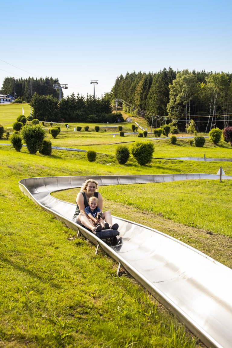 Frau rutscht mit ihrer Tochter auf einer Sommerrodelbahn hinunter, umgeben von Wiese und Wald