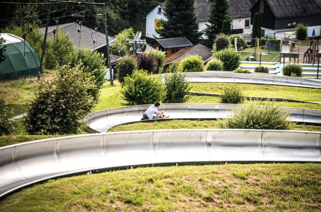 Man toboggans down a summer toboggan run, surrounded by forest and meadow